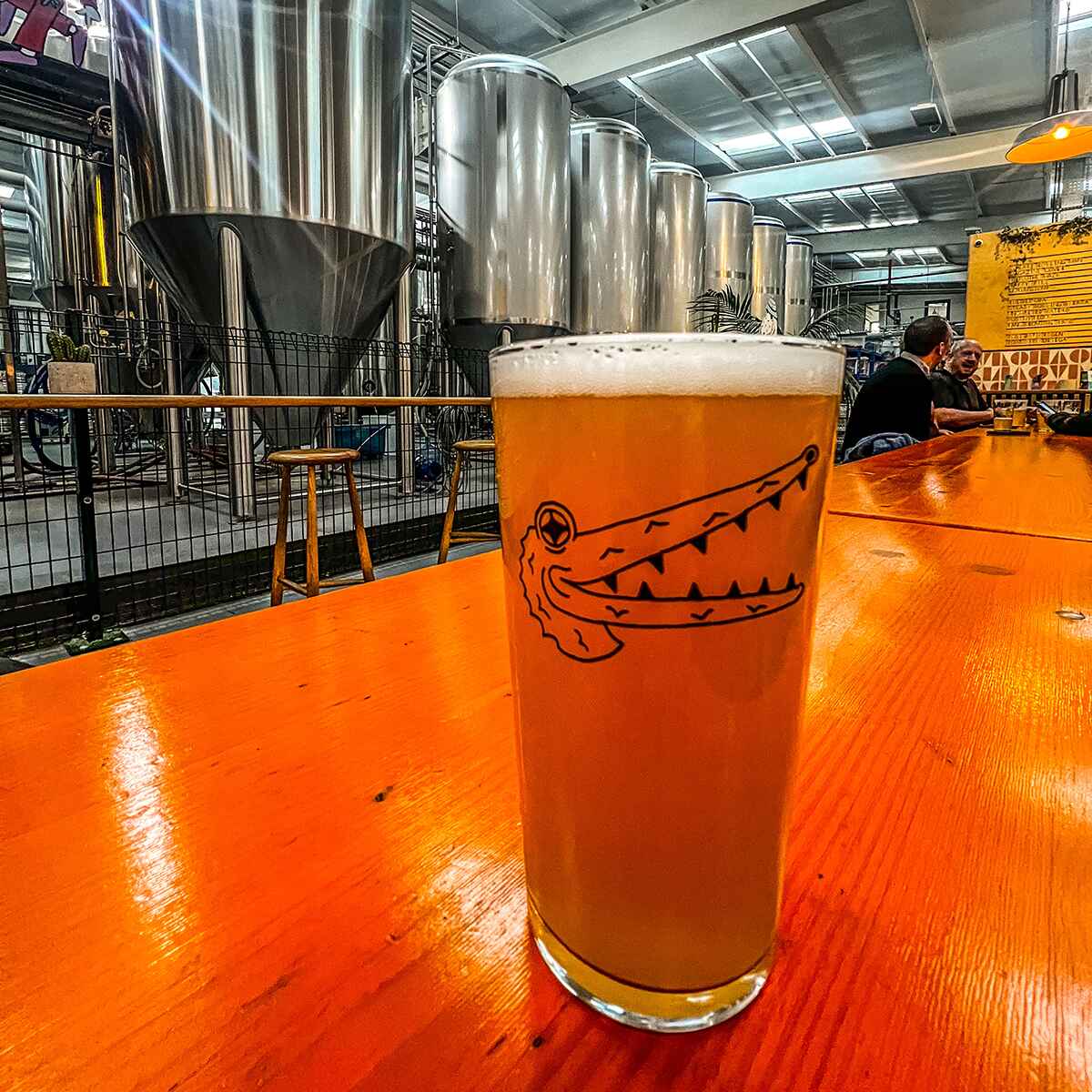 A pint of craft beer in a brewery with beer making equipment in the background. The glass has an unusual drawing of an unknown long nosed happy animal on it with sharp teeth.