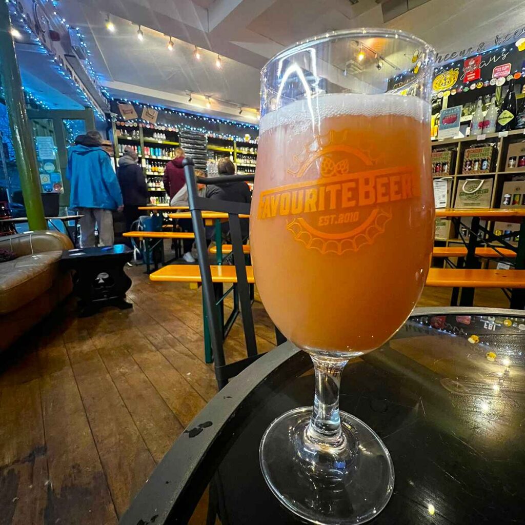 A glass of craft beer, with the inside of a specialist craft beer and cider off-licence in the background. The glass has the words 'favourite beer est 2010' written on it.