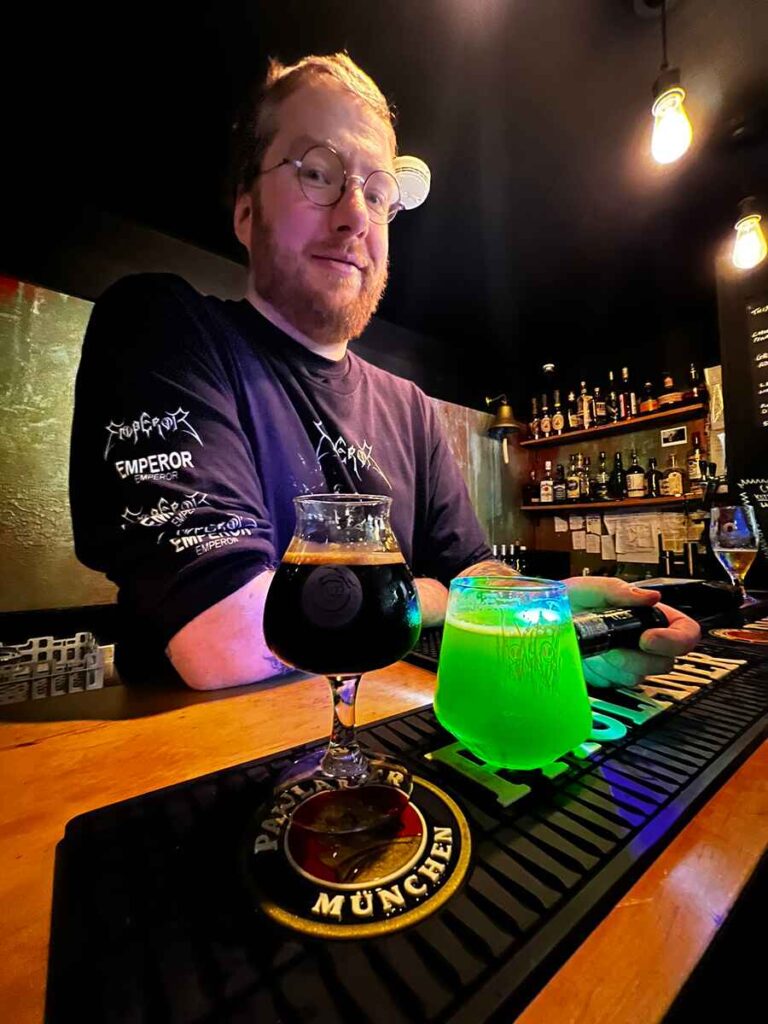 Bar tender smiling at bar with a glass of brown alcoholic drink and one shocking green drink