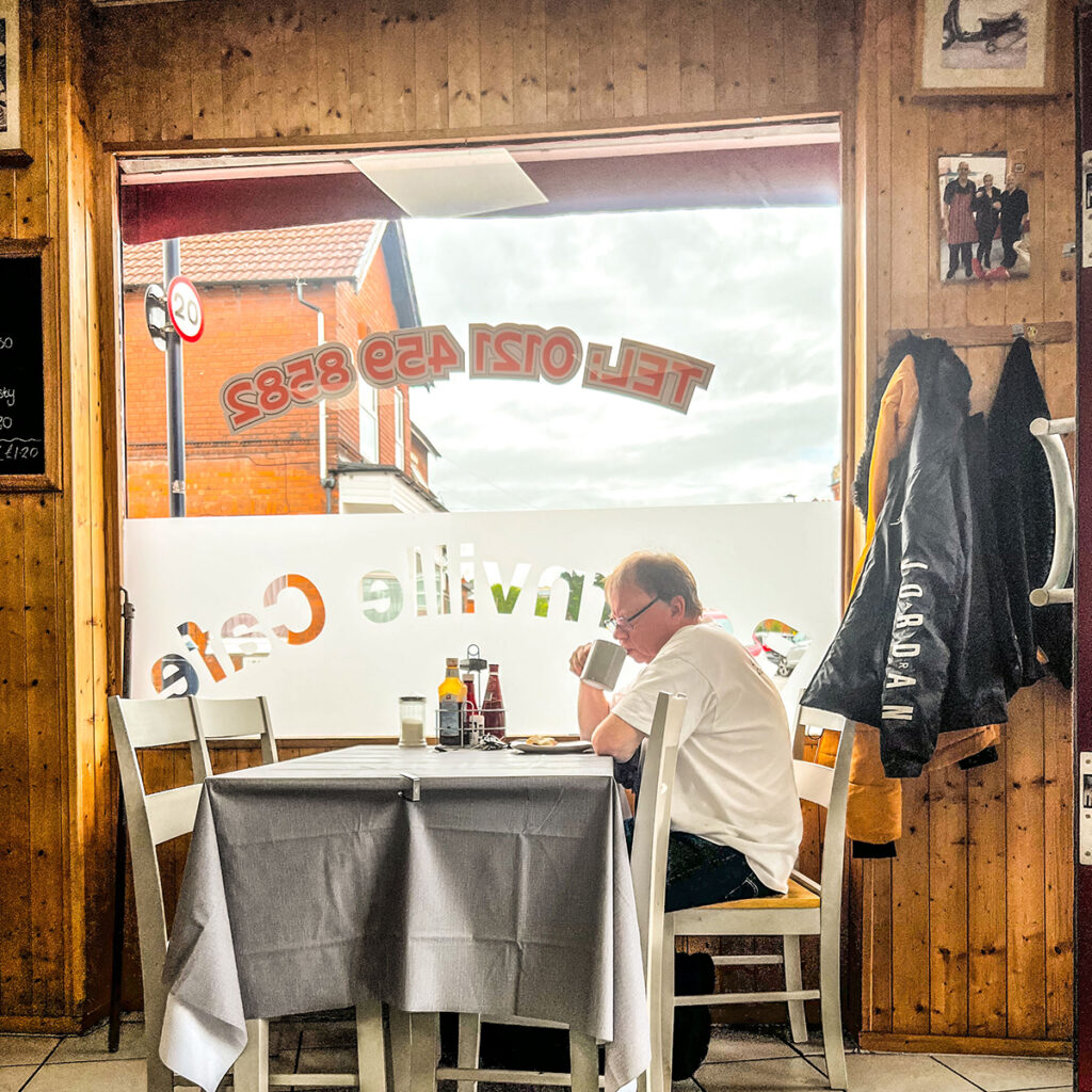 A person sips a mug of tea sat at a table next to the wood panelled interior of Bournville cafe.

