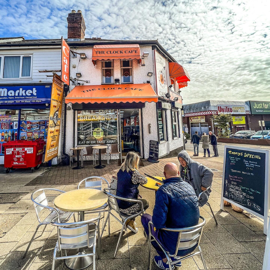 three people sat on table and chairs outside the front of the Clock Cafe in Northfield. 